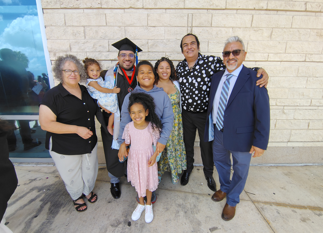 Ethan Galvan in cap and gown with medal poses with six family members, including two young children, outside a building. Oscra Macias is also in the group. Everyone is smiling in a group photo on a sidewalk.