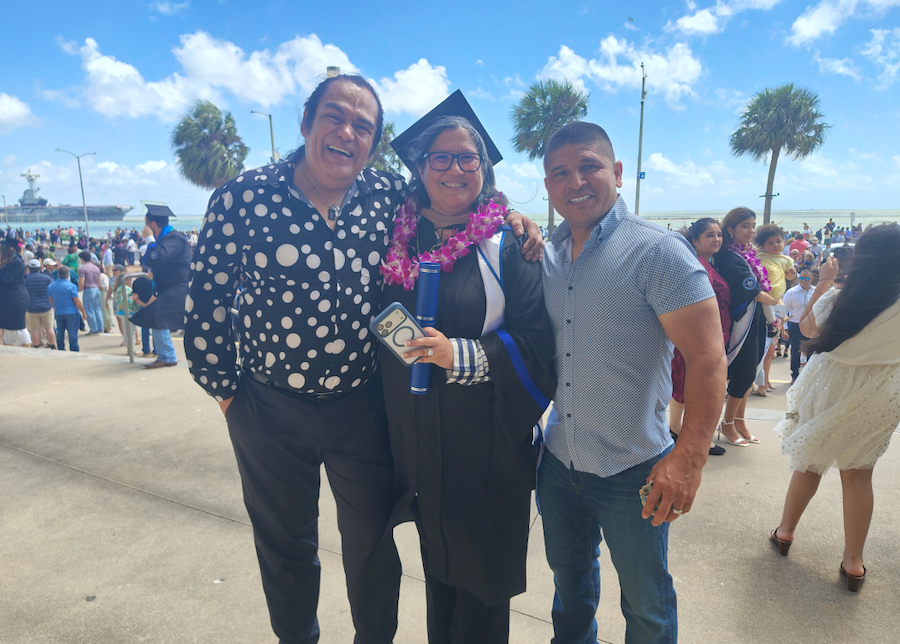 Melissa Lopez in cap and gown holds a diploma while flanked by Oscar Macias and another man outside near palm trees and a waterfront, with a crowd of graduation attendees in the background.