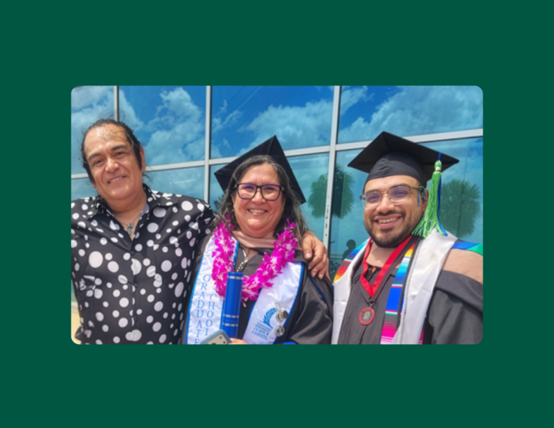 Two graduates in cap and gown pose with a Risepoint team member outside a glass building. The woman in the center wears a pink lei and holds a diploma; the man on the right wears a colorful serrape stole and medal. Both wear graduation regalia with green and blue tassels.