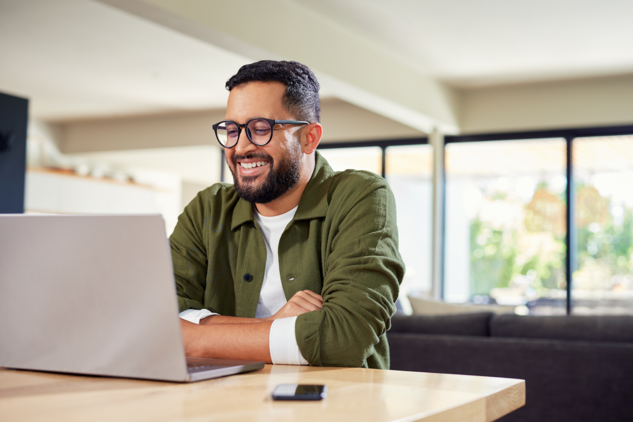 Smiling adult student with glasses studying on a laptop at a wooden table in a bright home living space.